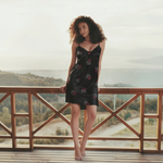 Woman in a black dress with floral patterns standing on a wooden deck with a scenic background.