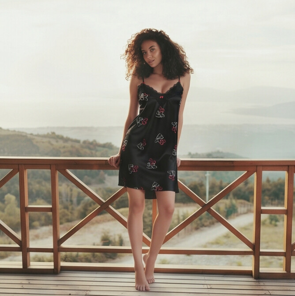 Woman in a black dress with floral patterns standing on a wooden deck with a scenic background.