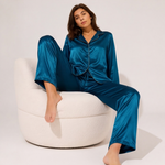 Woman wearing a sapphire blue satin pajama set featuring a long-sleeve button-down shirt with contrast piping and matching full-length satin pants.
