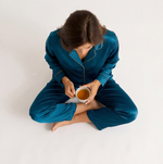 Woman wearing a sapphire blue satin pajama set featuring a long-sleeve button-down shirt with contrast piping and matching full-length satin pants.