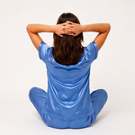Person wearing a blue satin pajama set sitting with hands behind head on a white background